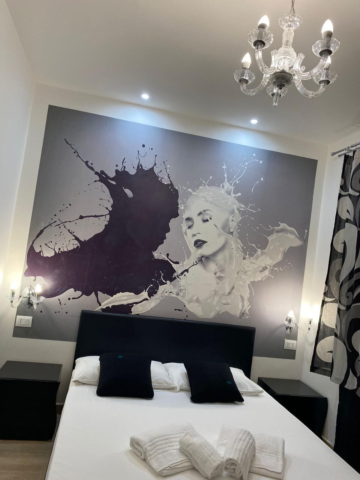 Une chambre d’hôtel moderne avec un grand tableau abstrait au mur, un lustre et des lits soigneusement faits, avec des serviettes à disposition.
