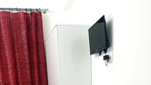 Une pièce avec un bureau blanc, deux chaises noires, un rideau rouge à motifs et un téléviseur fixé au mur.