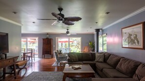 A spacious living room with a large brown sofa, a wooden coffee table, and a ceiling fan. There is a television on the left and a bookshelf in the background. - Lovely, Spacious Villa with HotTub, Outdoor Dining, Fire Pit (Volcano)