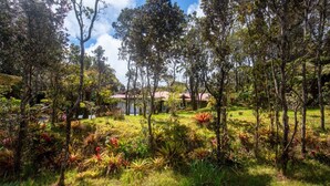 A lush garden with various plants and trees, a building with a red roof in the background, and a clear sky. - Lovely, Spacious Villa with HotTub, Outdoor Dining, Fire Pit (Volcano)