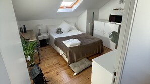 Habitació compacta i ben il·luminada amb sostre inclinat, un llit amb col·cha marró, una tauleta de nit, una làmpada, una petita planta i un radiador.