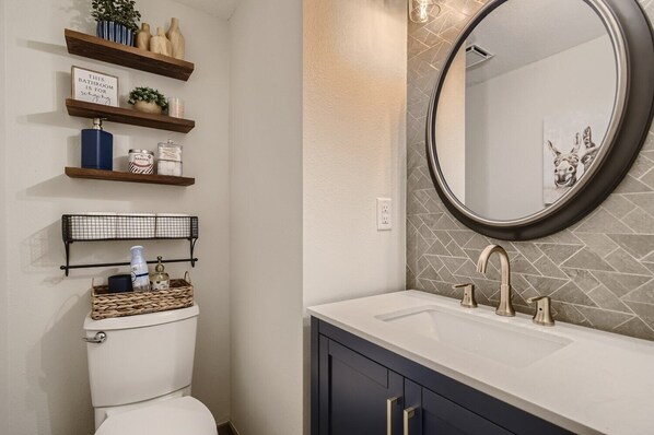 Un baño con inodoro blanco, tocador con un espejo grande y repisas sobre el inodoro que sostienen artículos de tocador y objetos decorativos.