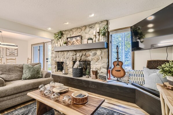 A cozy living room with a stone fireplace, a sofa, a coffee table with decorative items, and a guitar. - PET FRIENDLY 3-Bedroom Condo on the Creek! Newly Renovated! - CC206 (Frisco)