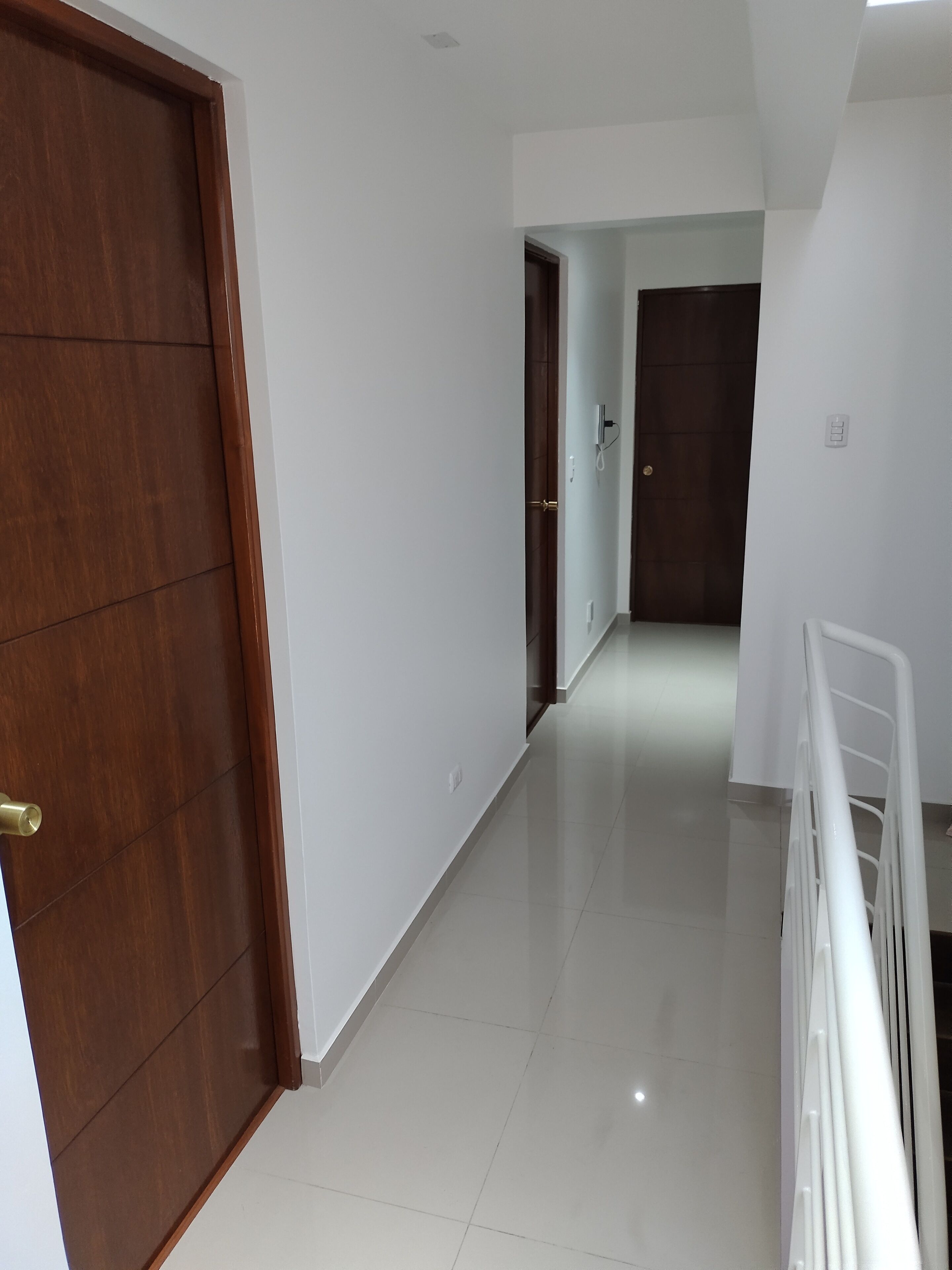 A hallway with a wooden door on the left, white walls, and a white railing on the right.