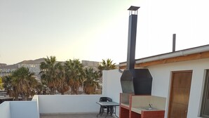 Una terraza en la azotea con zona de barbacoa, una mesa y sillas, y vistas a palmeras y edificios al fondo.
