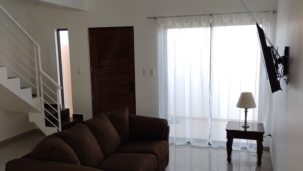 Un salón amplio con un sofá marrón, una escalera, una puerta de madera y un televisor montado en la pared.