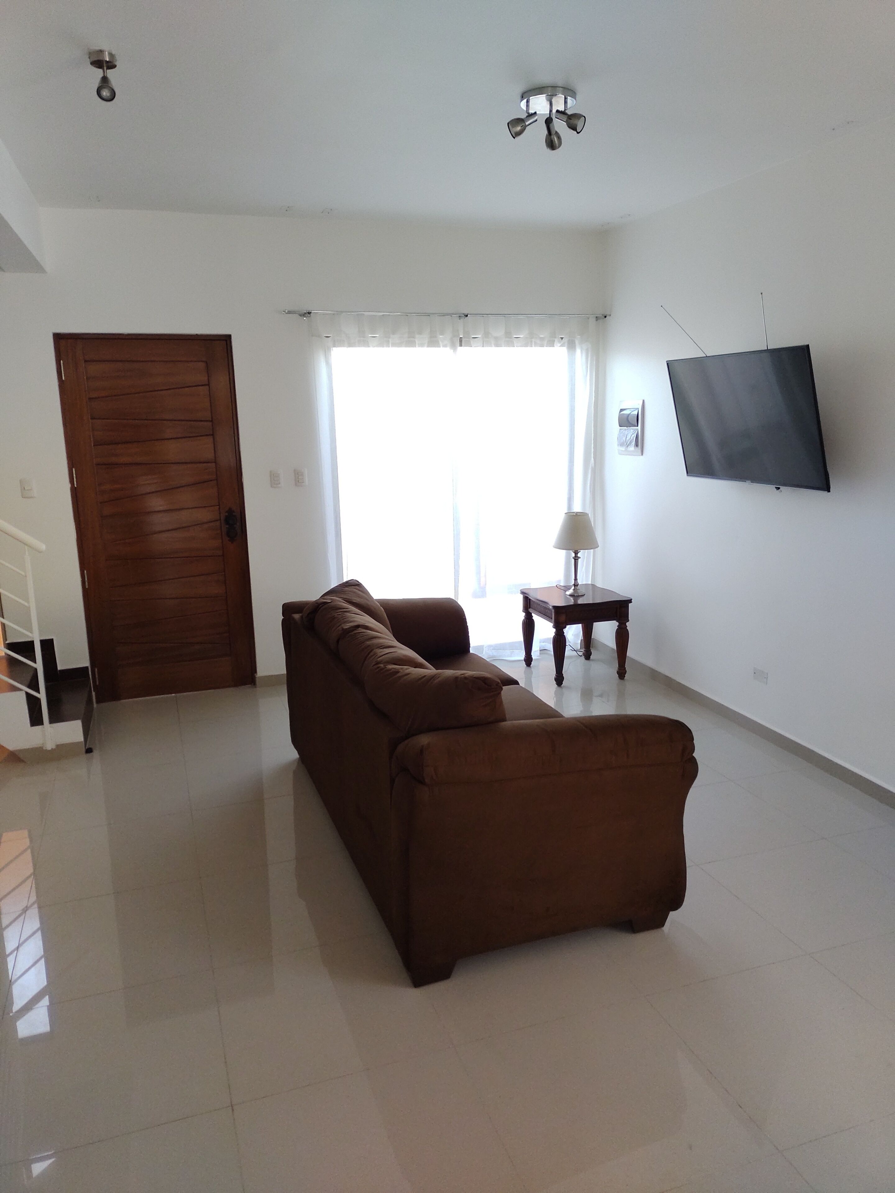 Un salón amplio con un sofá marrón, un televisor montado en la pared, una puerta de madera y una escalera que sube a un piso superior.