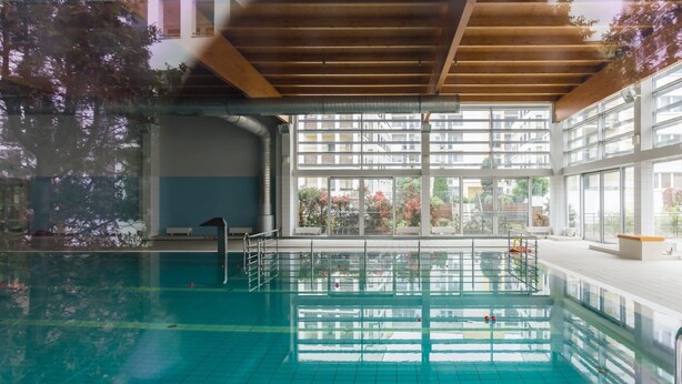 An indoor swimming pool with clear glass walls, wooden beams, and a view of a residential building. - Comfortable Apartment | Pool Access and Balcony (Poznań)