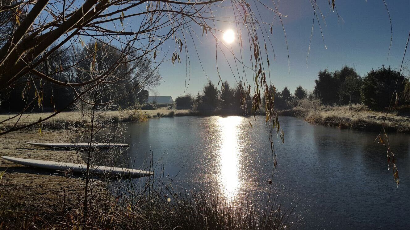Une scène paisible au bord de la rivière, où la lumière du soleil se reflète sur l’eau, entourée d’arbres et sous un ciel dégagé.