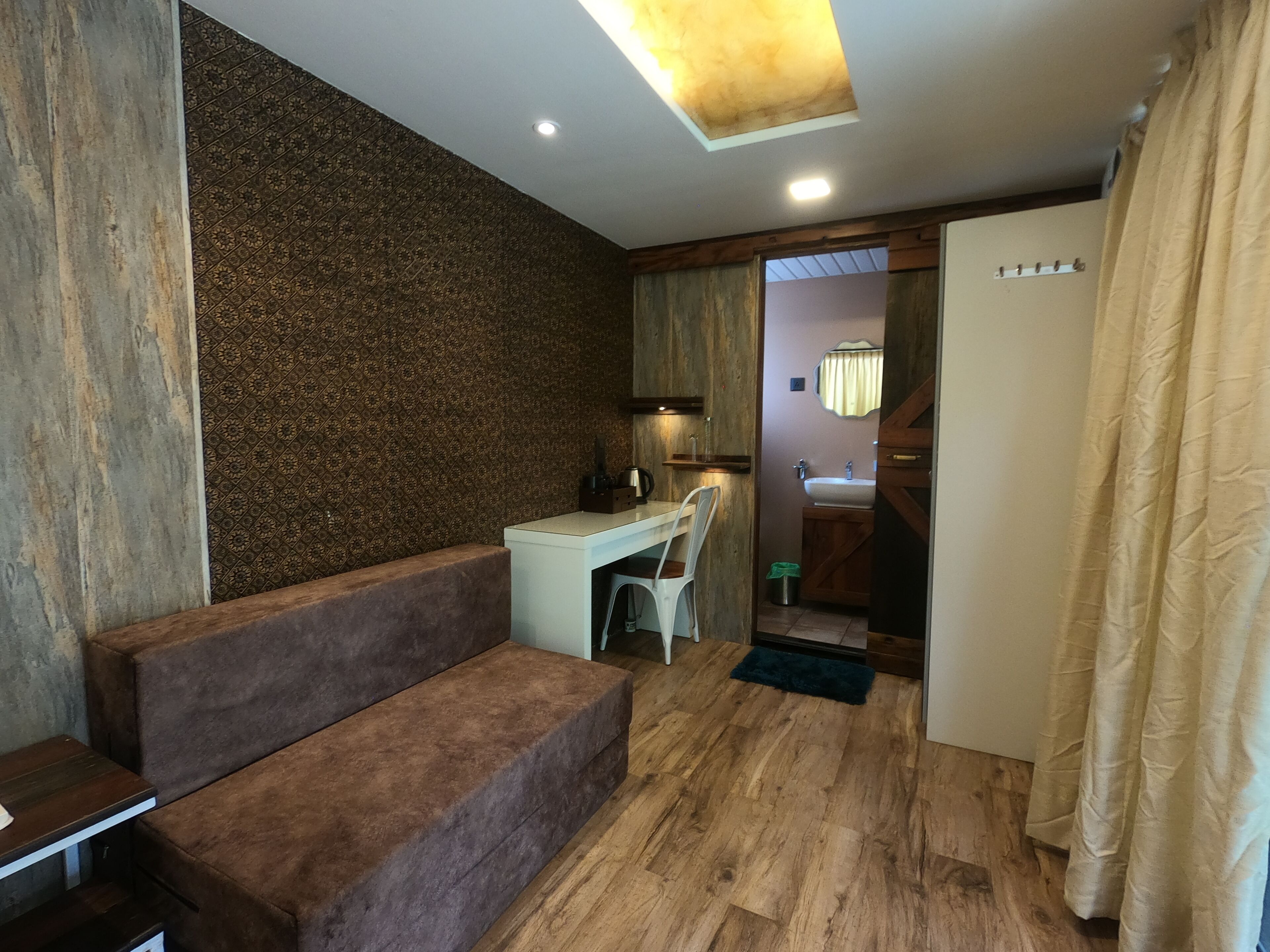 A compact room with a wooden floor, a brown sofa, a desk with a chair, a small trash bin, and a bathroom visible through an open door.