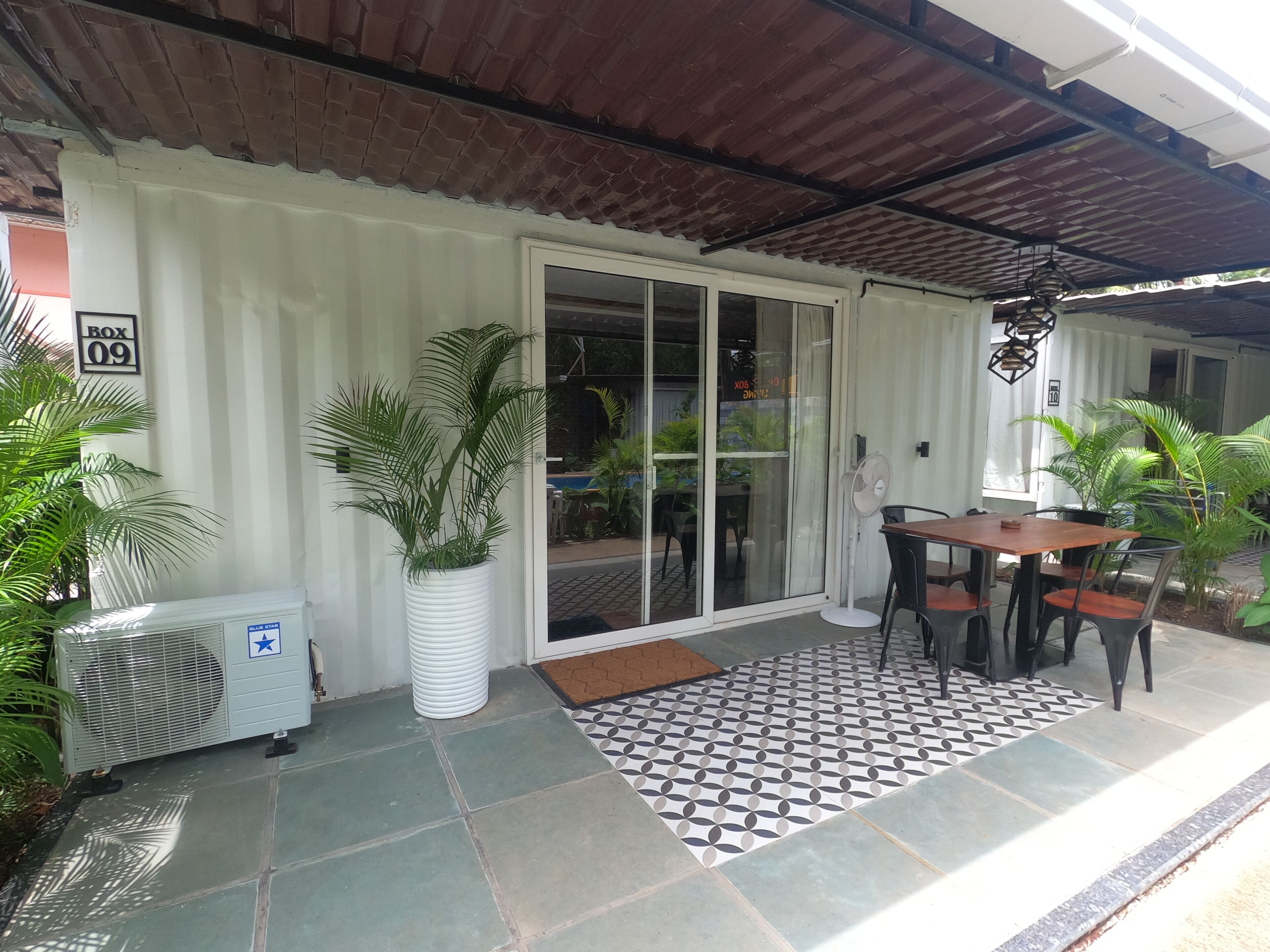 An outdoor patio area with a tiled floor, a table with chairs, a potted plant, and an air conditioning unit.