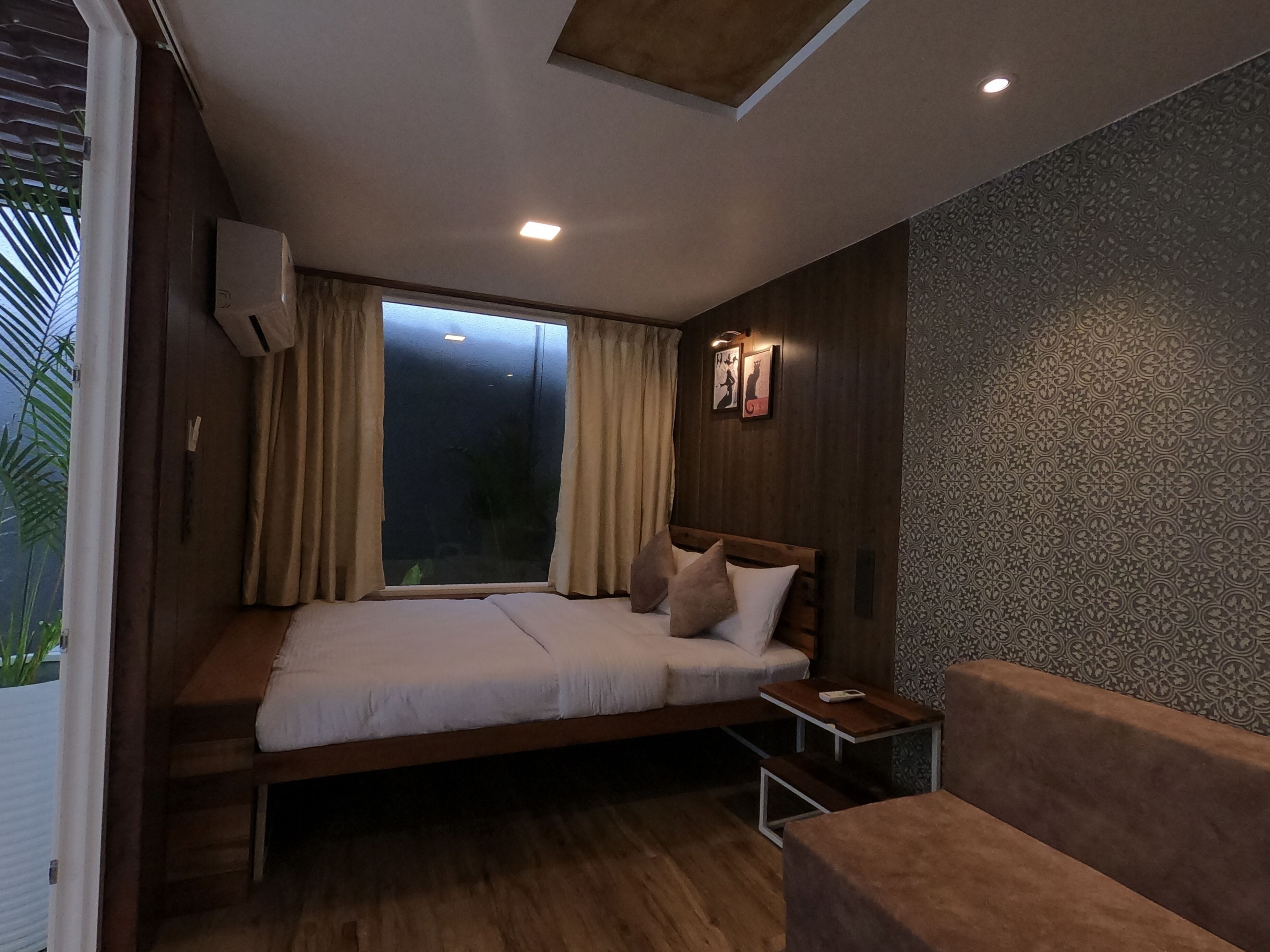 A bedroom with a bed, a nightstand, a sofa, a window with curtains, and a wall with a patterned design.