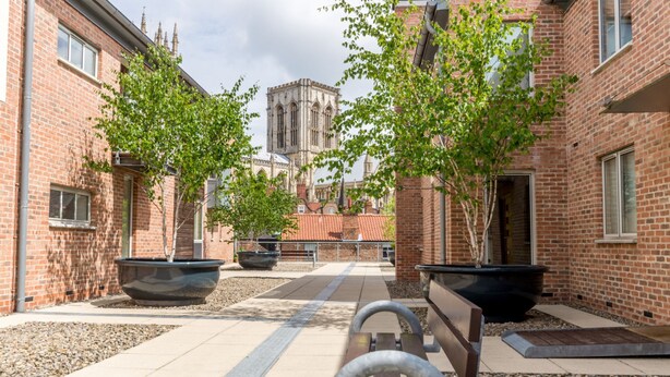 Une cour bordée de bâtiments en briques, de grands jardinières plantées d’arbres et d’un banc.