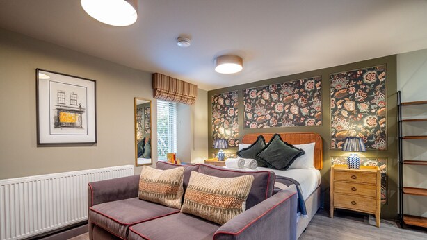 A bedroom with a bed, a sofa, a bedside table, and a wall with floral wallpaper. - The Falcon Apartment 6 (York)