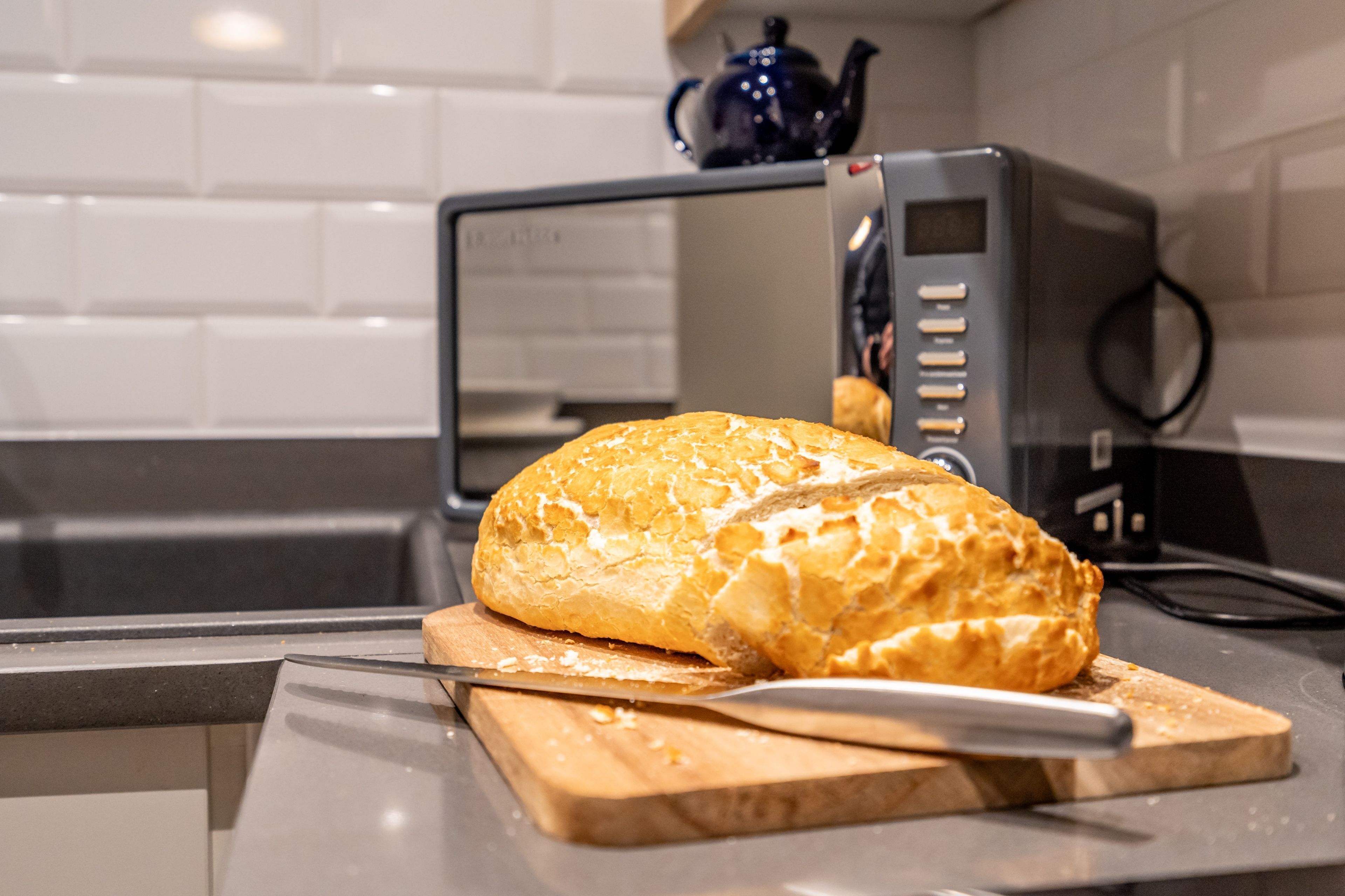 A loaf of bread on a cutting board with a knife, in a kitchen with a microwave and a kettle.