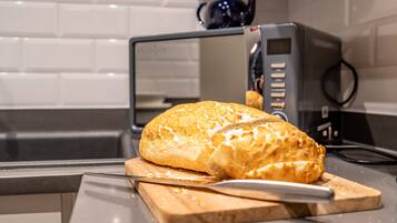 A loaf of bread on a cutting board with a knife, in a kitchen with a microwave and a kettle.