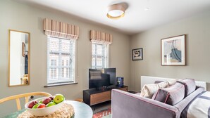 A modern living room with a flat-screen TV, a sofa, a dining table with a bowl of fruit, and large windows with striped valances. - Studio - Sleeps 2 Guests (York)