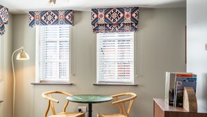 A room with two windows, a round table, two wicker chairs, a floor lamp, and a wooden sideboard with books. - The Falcon Tap Apartment 2 (York)