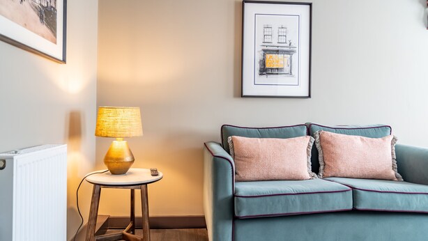 A living room with a teal sofa, two cushions, a lamp on a wooden stool, and a framed picture on the wall. - The Falcon Tap Apartment 2 (York)
