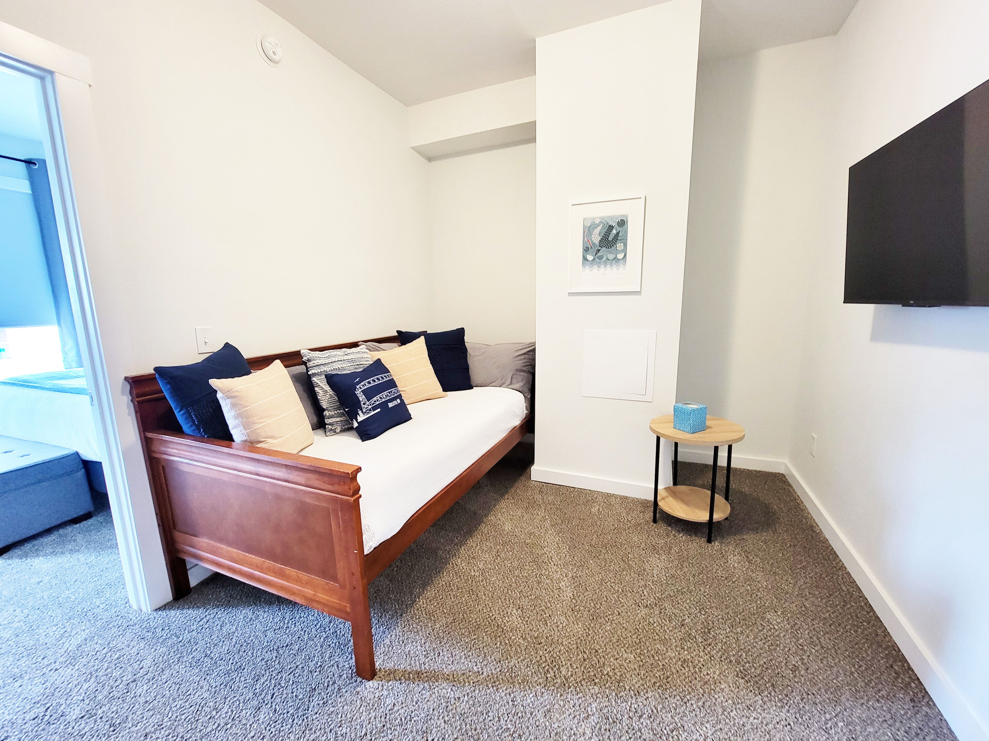 Une chambre avec un lit en bois, une petite table ronde, un téléviseur fixé au mur et une porte donnant sur une autre pièce.