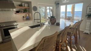 Cozinha moderna com ilha central, armários brancos, eletrodomésticos de aço inoxidável e vista para o oceano.