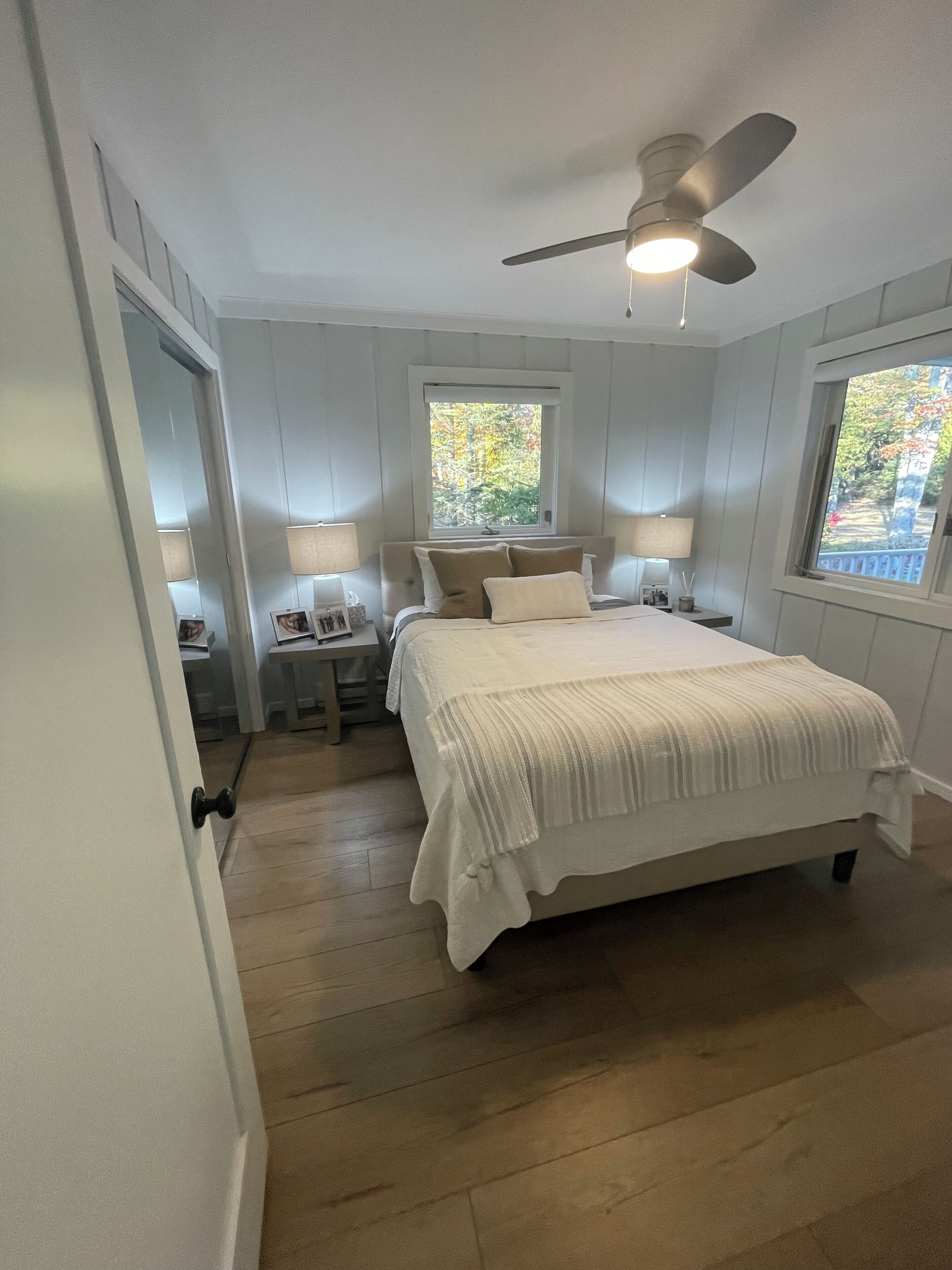 A bedroom with a bed, two bedside tables, a ceiling fan, and windows with a view of trees.