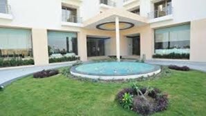 A modern building with a central courtyard featuring a circular water feature and surrounding greenery. - KRSNA Lila By The Blues Hotels (Sanand)