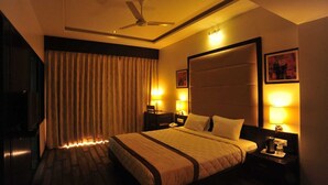 A hotel room with a bed, two bedside lamps, a ceiling fan, and a large window with curtains. - KRSNA Lila By The Blues Hotels (Sanand)