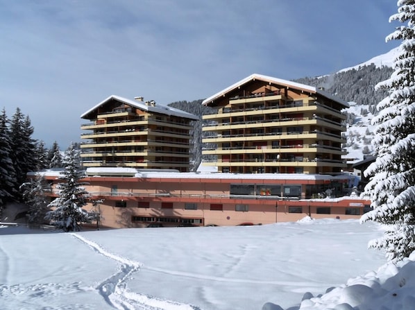 Un hotel de varios pisos con balcones, ubicado en un paisaje nevado con montañas al fondo.