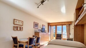A hotel room with a bed, a desk with a computer, chairs, and framed pictures on the wall. - Studio *** - 6th floor - close to the centre, shops and bus stop - sunny balcony with very nice view (Verbier)