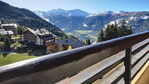 A mountain village with traditional wooden houses, a wooden balcony railing, and a valley with snow-capped mountains in the background. - Studio *** - 6th floor - close to the centre, shops and bus stop - sunny balcony with very nice view (Verbier)