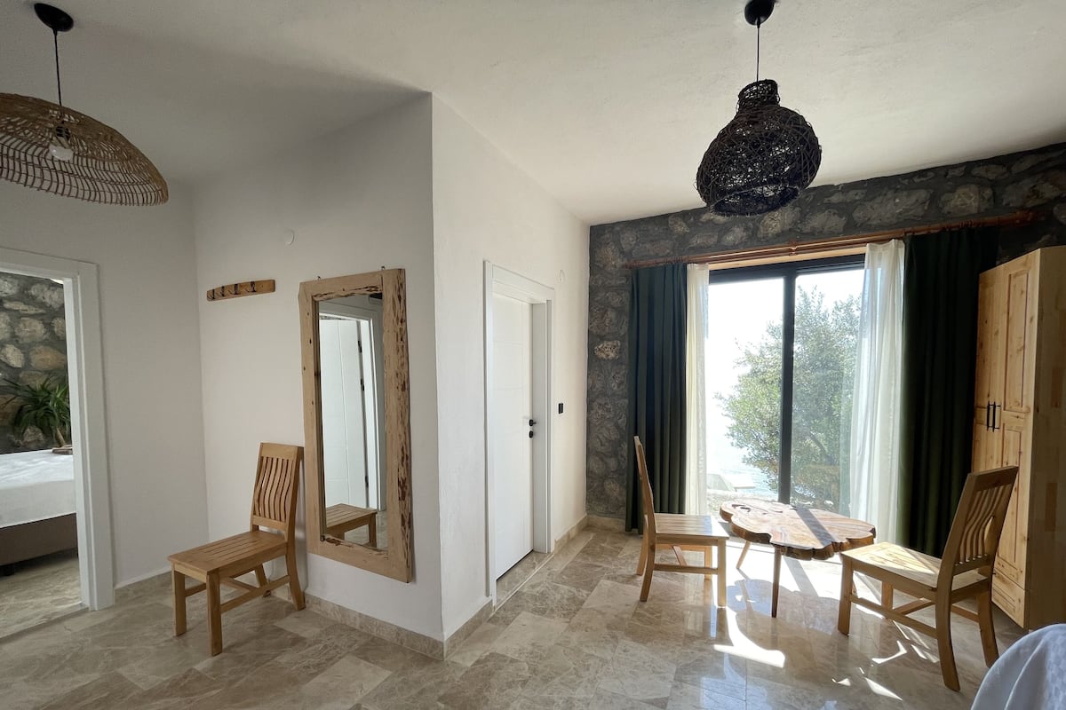 Une pièce spacieuse avec un mur en pierre, un grand miroir, des chaises en bois, une table ronde et une vue sur l’extérieur, visible à travers une porte vitrée.
