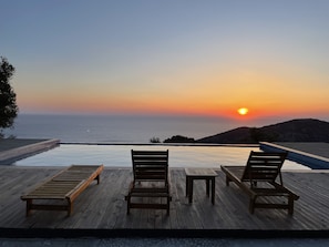 Een houten terras met drie loungestoelen en een kleine tafel, met uitzicht op een zwembad en de oceaan bij zonsondergang.