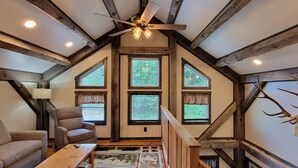 Une pièce chaleureuse avec un plafond en pente, des poutres en bois, un ventilateur de plafond et une grande fenêtre avec des rideaux.