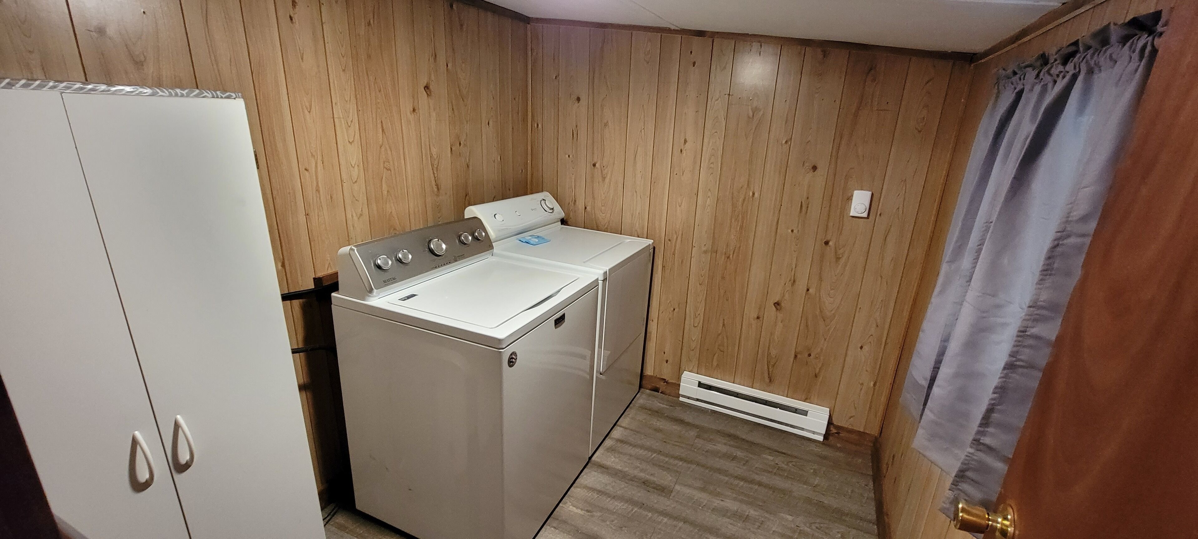 A laundry room with a white washer and dryer, wooden walls, a white cabinet, and a vent.