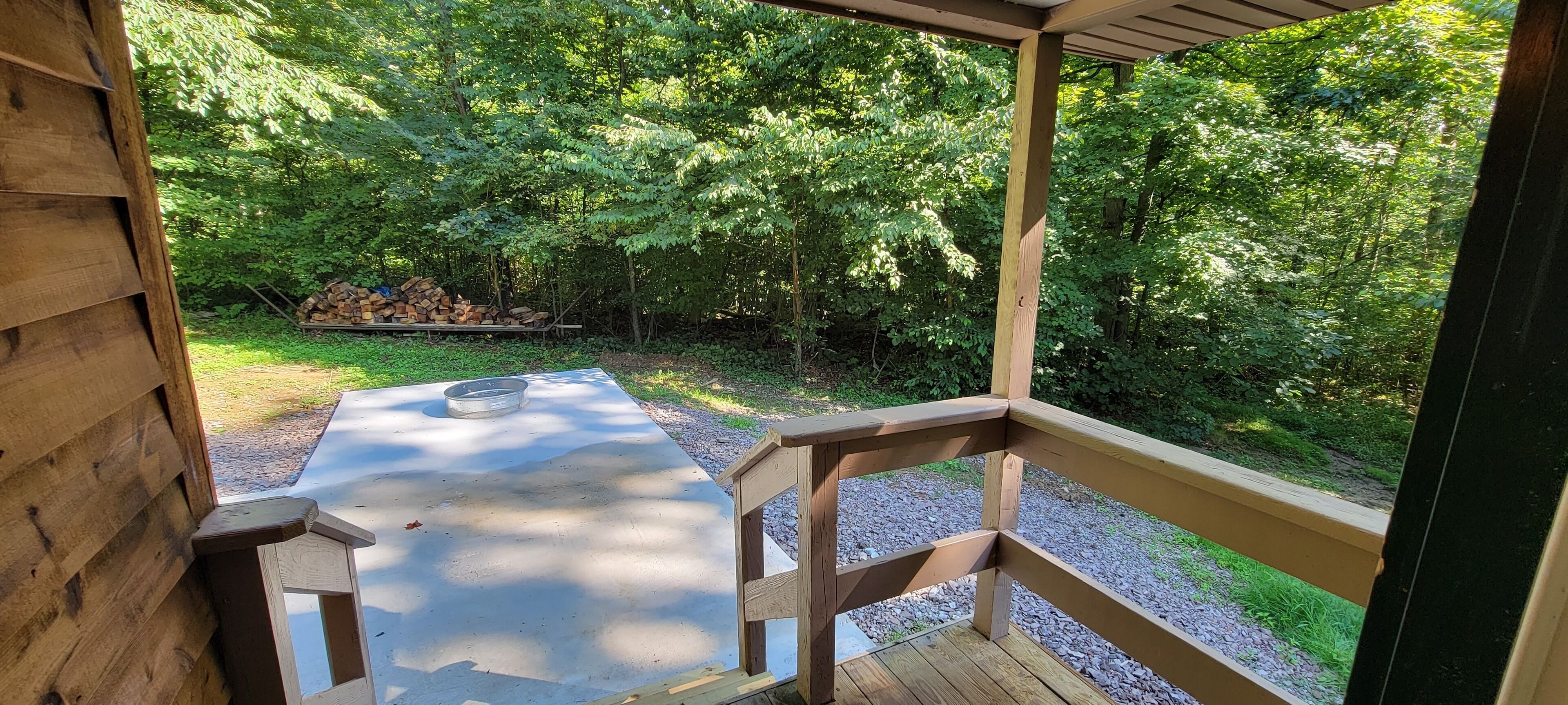 A covered outdoor area with a concrete floor, wooden railings, and a view of a forested area.