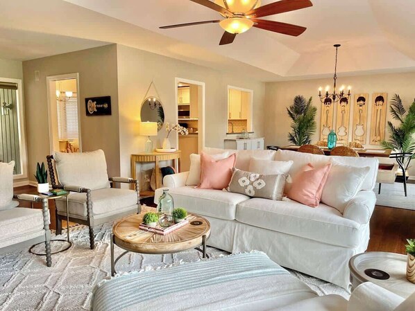 A living room with a white sofa, a wooden coffee table, a ceiling fan, and a kitchen visible in the background.