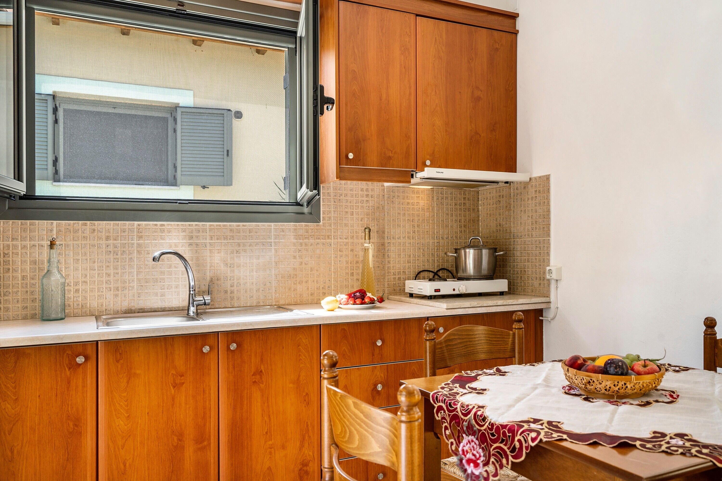 Une cuisine avec des meubles de rangement en bois, un évier, une fenêtre, une cuisinière et une table sur laquelle se trouve un panier de fruits.