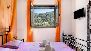 Un letto con copripiumino floreale viola, una finestra con tende arancioni e vista su un paesaggio verde.