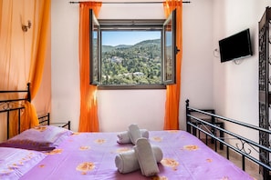 A bed with a purple floral bedspread, a window with orange curtains, and a view of a green landscape.