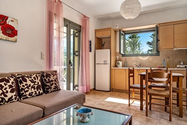 A modern kitchen with a refrigerator, wooden cabinets, a dining table with chairs, and a sofa with patterned cushions. - Holiday Accommodation "Alkionides Apartment 1" with pine forest overlook (Lefkada)