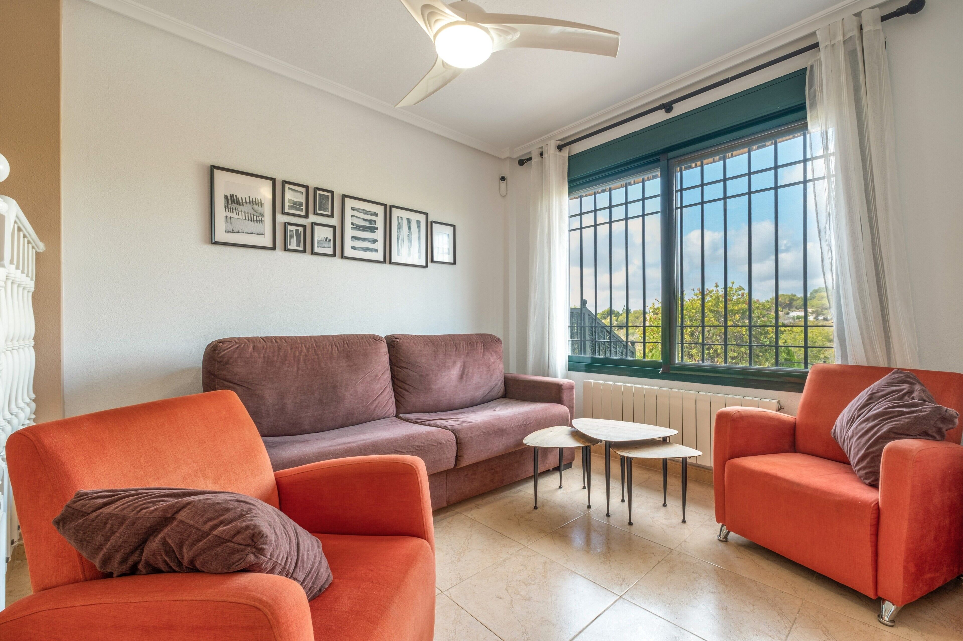 Ein Wohnzimmer mit einem braunen Sofa, zwei orangefarbenen Sesseln, einem kleinen weißen Tisch und einem Fenster mit Blick auf Grünflächen.