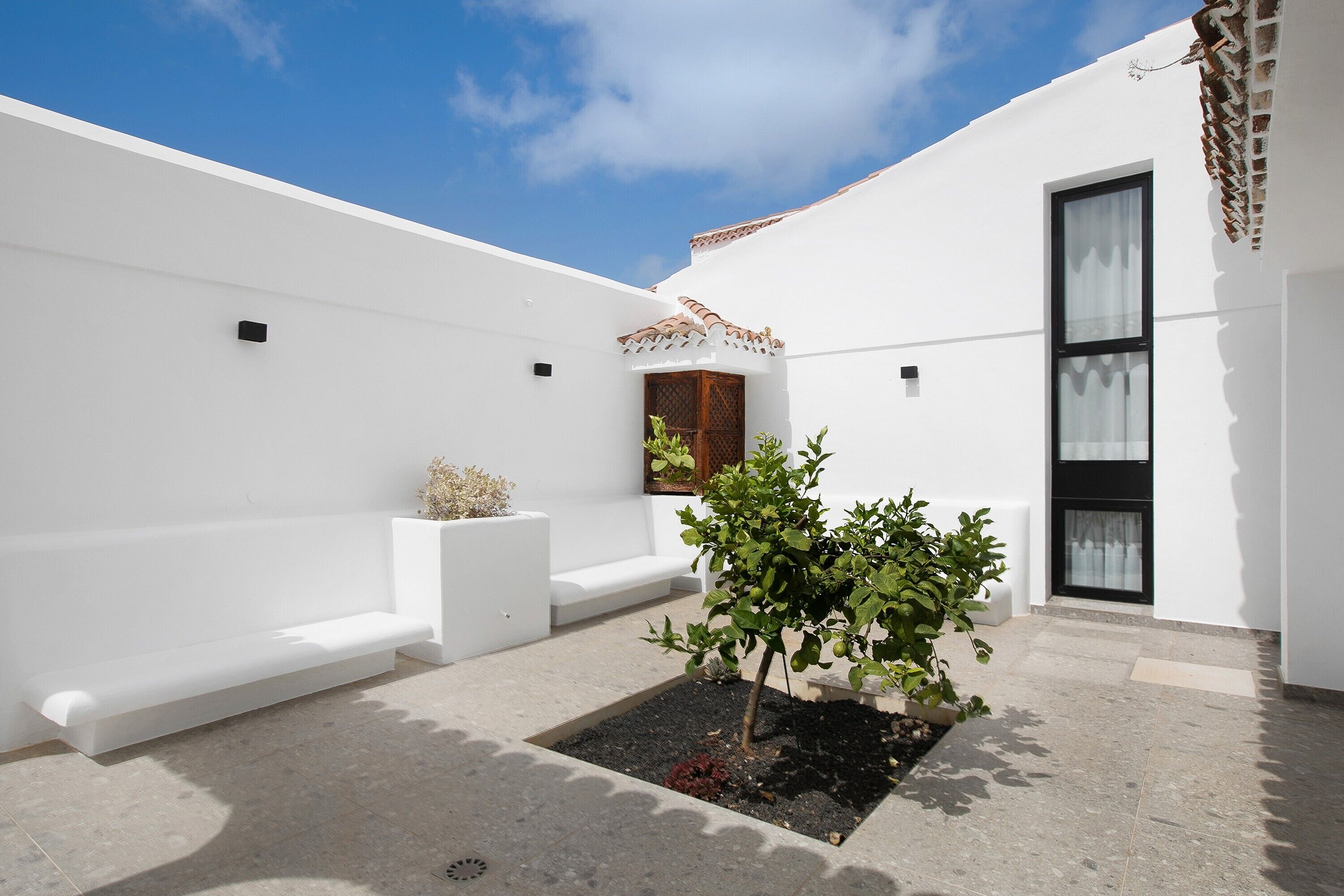A white building with a tiled roof, a small garden area with a tree, and a bench.