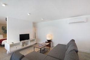 A modern living room with a sofa, a TV on a white stand, a lamp, and a small table with books.