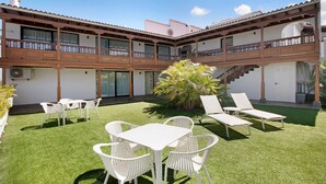 Un patio con una mesa y sillas blancas, una pequeña palmera y un edificio con balcones y una escalera.