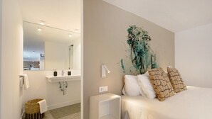 Una habitación de hotel moderna con una cama grande, una mesita de noche, una lámpara de pared, un baño con lavamanos y espejo, y un adorno en la pared.