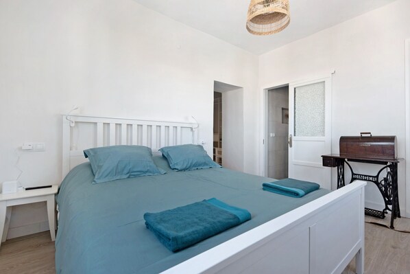 A bedroom with a white bed, a nightstand, a wicker hanging light, and a Singer sewing machine on a table.