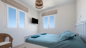 Een slaapkamer met een bed, twee ramen, een televisie, een rieten stoel en een hanglamp.