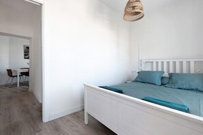 A bedroom with a white bed, teal bedding, a wicker hanging light, and a view of a dining area with a table and chairs.
