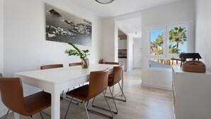 Een moderne eetruimte met een witte tafel, bruine stoelen, een zwart-wit kunstwerk met een kustlandschap en uitzicht op palmbomen door een raam.